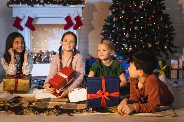 Happy multiethnic kids holding presents on blanket near fireplace and christmas tree at home — Stock Photo