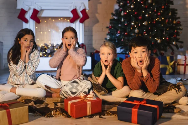 Niños multiétnicos asustados mirando la cámara cerca de regalos y el árbol de Navidad borroso en casa por la noche - foto de stock