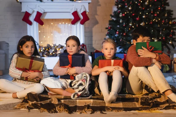 Des enfants interraciaux effrayés tenant des cadeaux près d'une cheminée floue et d'un arbre de Noël à la maison — Photo de stock