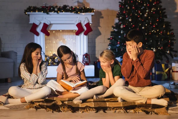 Chica leyendo libro cerca asustado interracial amigos en manta cerca de árbol de Navidad en casa - foto de stock