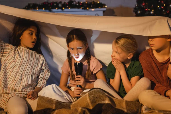 Chica sosteniendo la linterna cerca de amigos asustados bajo la manta durante la Navidad en casa - foto de stock