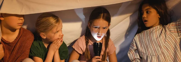 Mädchen hält Taschenlampe neben verängstigten Freunden unter Decke zu Hause, Banner — Stockfoto