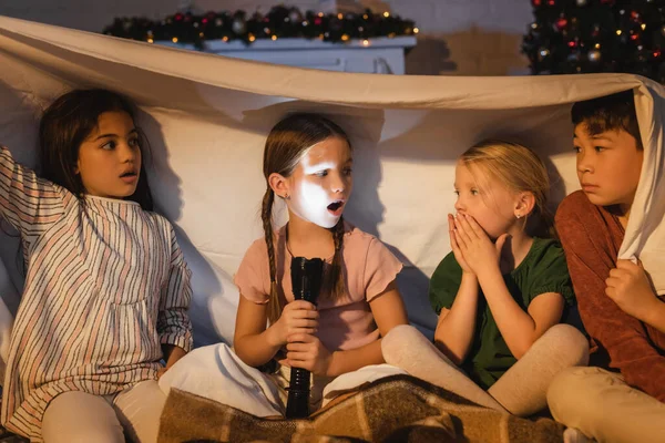 Chica sosteniendo linterna cerca asustado interracial amigos bajo manta y navidad decoración en casa - foto de stock