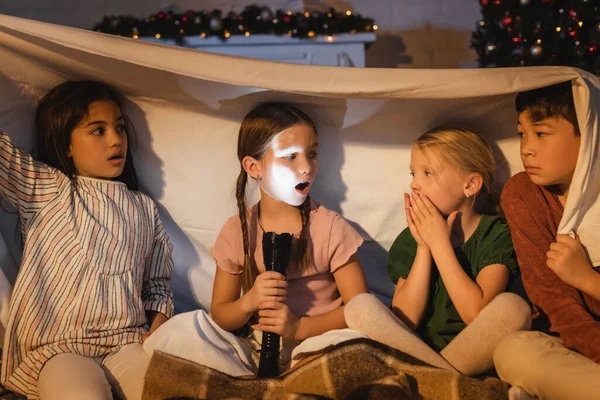 Menina segurando lanterna perto assustados amigos inter-raciais sob cobertor e decoração de Natal em casa — Fotografia de Stock