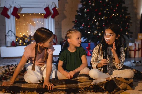 Chica con linterna hablando cerca de amigos en manta y borrosa árbol de Navidad en casa - foto de stock