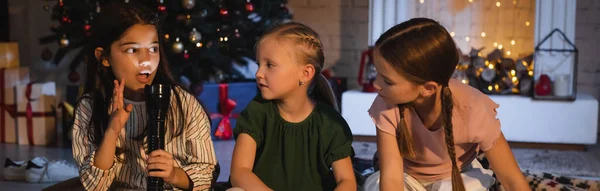Kid holding flashlight and talking to friends near blurred christmas decor at home, banner — Stock Photo