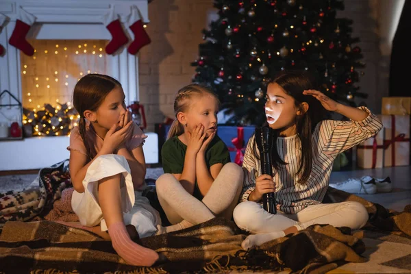 Preteen girl holding flashlight and talking to scared friends on blanket near blurred fireplace with christmas decor — Stock Photo