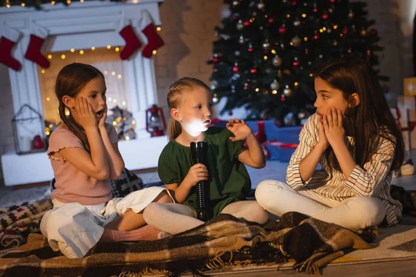 Child with flashlight talking near afraid friends on blanket during christmas celebration at home — Stock Photo
