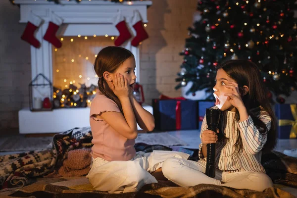 Ragazza con torcia sorridente vicino amico spaventato sulla coperta vicino sfocato albero di Natale a casa — Foto stock