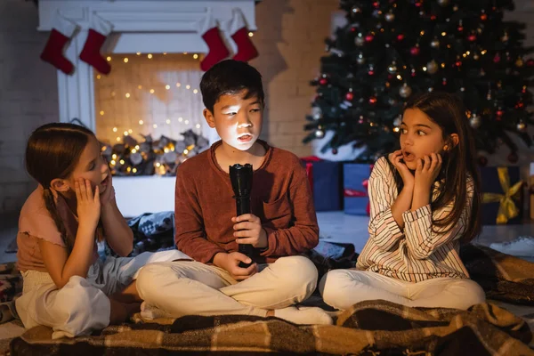 Chico asiático con linterna hablando cerca de niños asustados durante la celebración de Navidad en casa - foto de stock