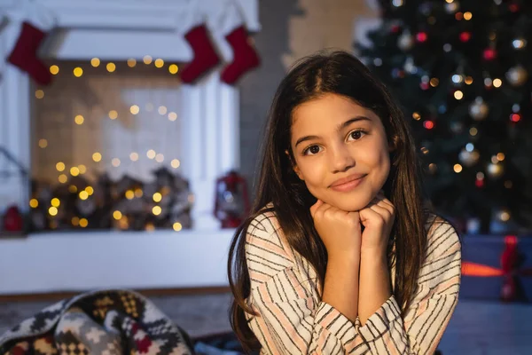 Cheerful girl looking at camera near blurred fireplace with christmas decor at home in evening — Stock Photo