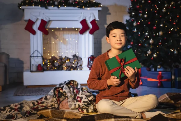 Asian boy holding gift box while sitting near blurred christmas tree at home — Stock Photo