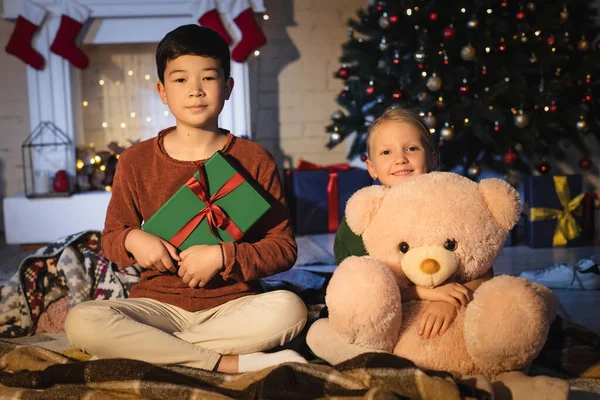 Asiático chico sosteniendo presente cerca amigo con suave juguete y árbol de Navidad en casa - foto de stock