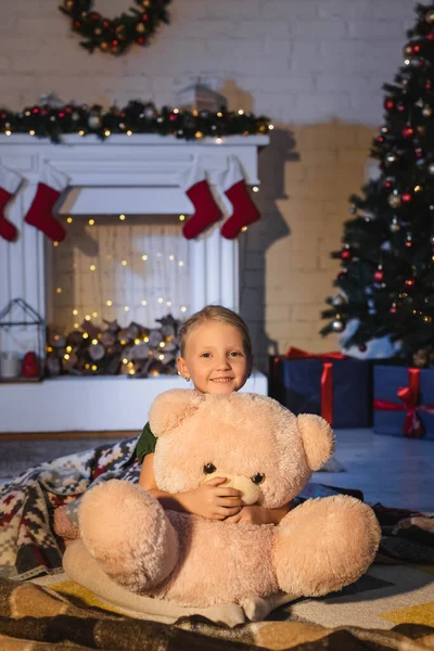 Niño sonriendo y abrazando juguete suave cerca de la chimenea borrosa y el árbol de Navidad en casa por la noche - foto de stock