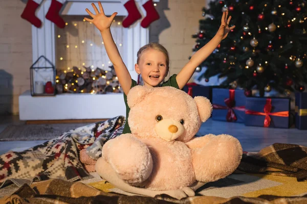 Menina positiva olhando para a câmera perto de ursinho de pelúcia em cobertor e árvore de natal borrada em casa — Fotografia de Stock