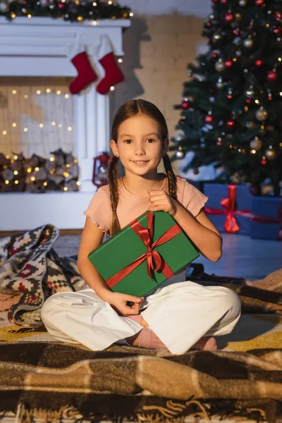 Preteen child holding present while sitting on blanket near christmas tree and fireplace at home — Stock Photo