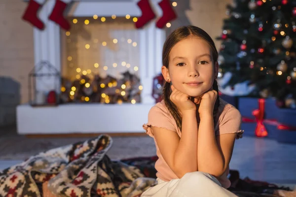 Enfant regardant la caméra près de couverture et décor de Noël flou à la maison le soir — Photo de stock