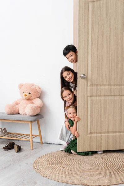 Щасливі багатоетнічні діти дивляться на камеру біля відкритих дверей в коридорі вдома — стокове фото