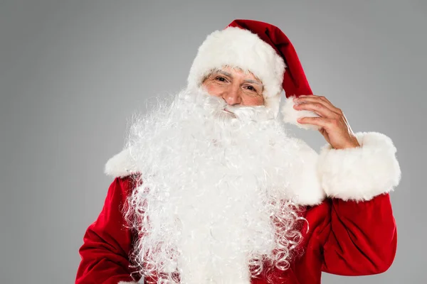 Père Noël en costume touchant chapeau et regardant la caméra isolée sur gris — Photo de stock