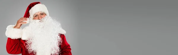 Père Noël en costume touchant chapeau et regardant la caméra isolée sur gris, bannière — Photo de stock