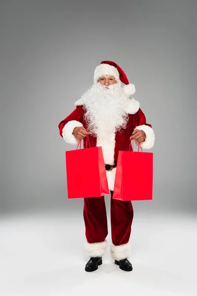 Санта-Клаус в длину держит красные сумки для покупок на сером фоне — стоковое фото