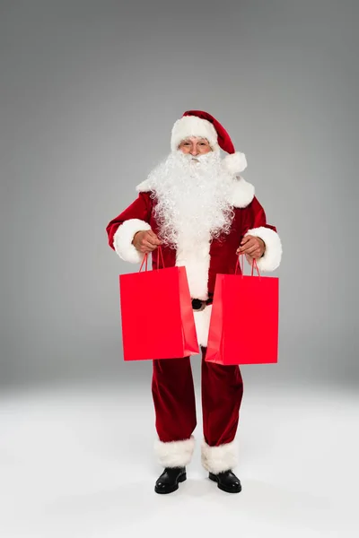 Pleine longueur de santa claus tenant des sacs à provisions rouges sur fond gris — Photo de stock