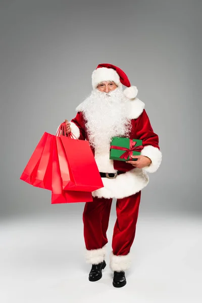 Pleine longueur de santa claus tenant des sacs à provisions rouges et cadeau sur fond gris — Photo de stock