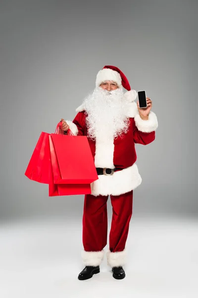 Weihnachtsmann im Kostüm hält Einkaufstüten und Smartphone mit leerem Bildschirm auf grauem Hintergrund — Stockfoto