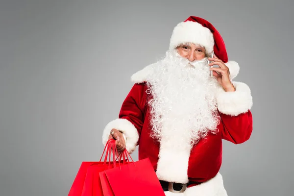 Weihnachtsmann telefoniert mit Smartphone und hält Einkaufstüten isoliert in Grau — Stockfoto