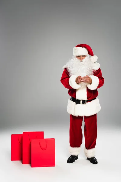 Toute la longueur du père Noël en utilisant un téléphone portable près des sacs à provisions sur fond gris — Photo de stock