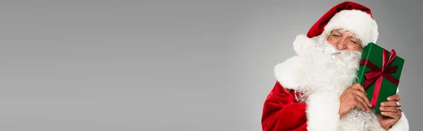 Father christmas in hat closing eyes while holding gift with bow isolated on grey, banner — Stock Photo