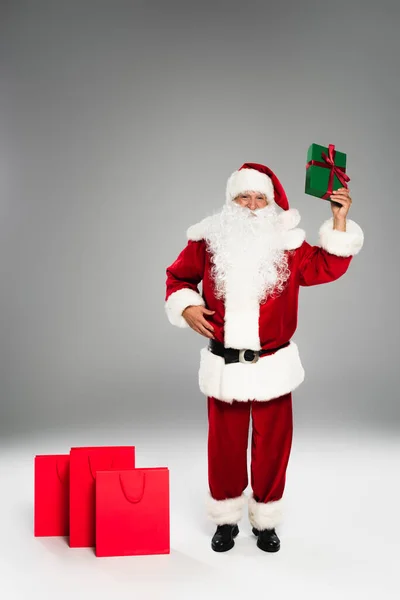 Повна довжина Санта-Клауса тримає подарункову коробку біля червоних сумки для покупок на сірому фоні — стокове фото