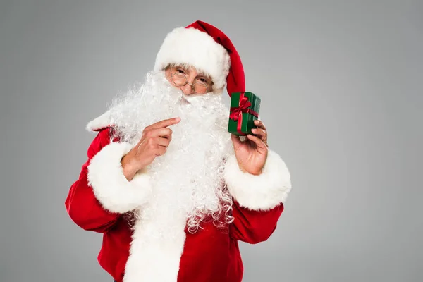 Père Noël dans des lunettes pointant vers une petite boîte cadeau isolée sur gris — Photo de stock