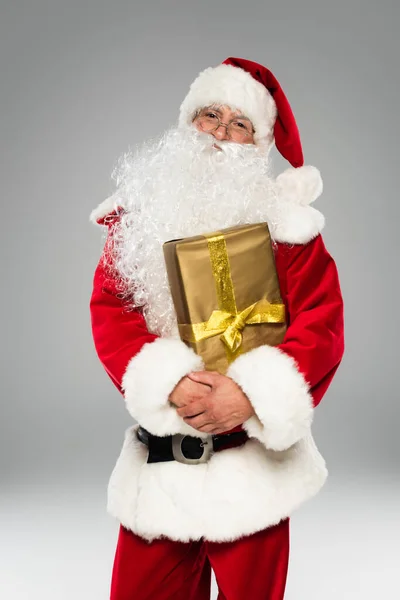 Санта-Клаус в шляпе и очках, держащий подарок и смотрящий на камеру, изолированную на сером — стоковое фото