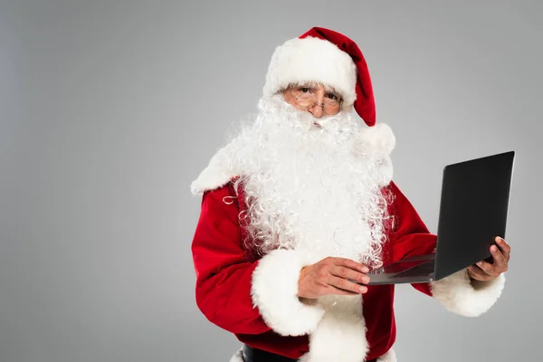 Санта-Клаус в окулярах і костюмах тримає ноутбук ізольовано на сірому — стокове фото