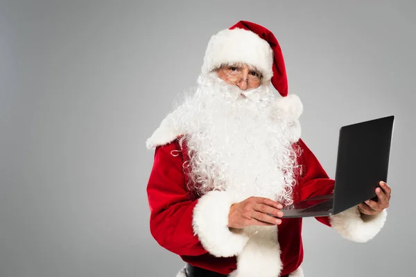 Santa claus in eyeglasses and costume holding laptop isolated on grey — Stock Photo