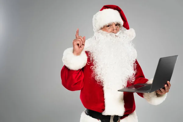 Санта-Клаус в очках, имеющий идею и держащий ноутбук изолирован на сером — стоковое фото