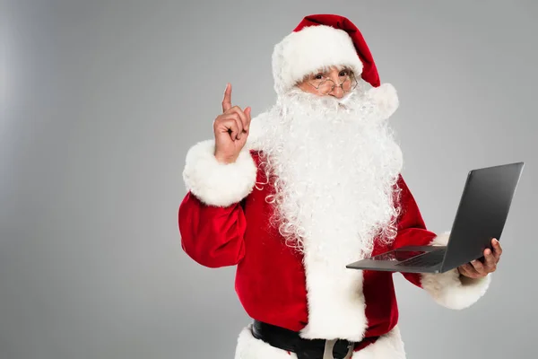 Santa claus in eyeglasses having idea and holding laptop isolated on grey — Stock Photo