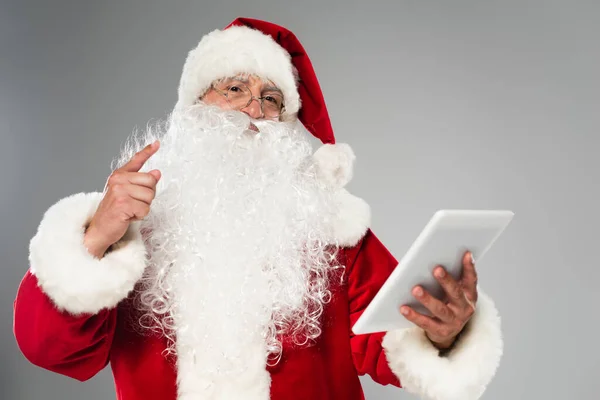 Батько Різдво в окулярах вказує пальцем і тримає цифровий планшет ізольовано на сірому — стокове фото