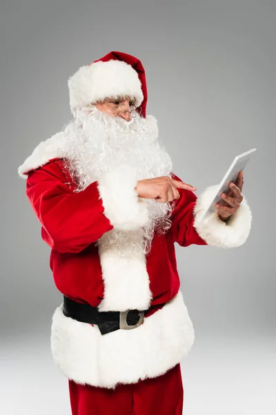 Санта-Клаус в окулярах, вказуючи на цифровий планшет ізольовано на сірому — стокове фото