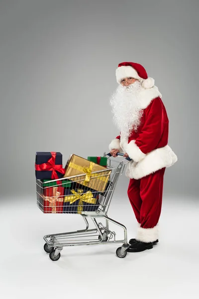 Longitud completa de Santa Claus en traje y gafas de pie cerca del carrito de la compra con regalos sobre fondo gris - foto de stock