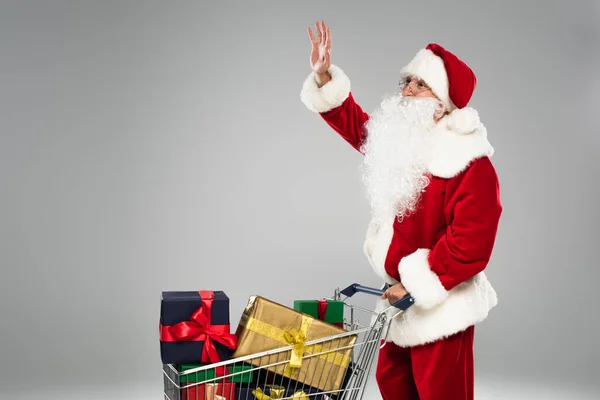 Santa Claus en sombrero agitando la mano cerca del carrito de la compra con regalos aislados en gris - foto de stock