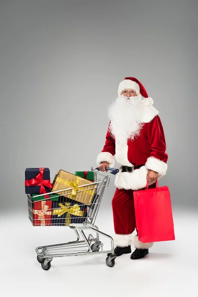 Babbo Natale tiene borse della spesa vicino al carrello con regali su sfondo grigio — Foto stock