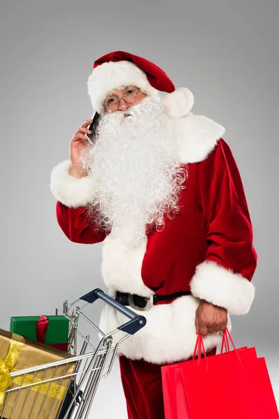 Santa claus in eyeglasses talking on smartphone and holding shopping bags near cart with gifts isolated on grey — Stock Photo