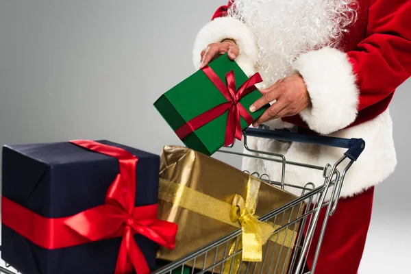 Cropped view of santa claus in costume putting gift box in shopping cart isolated on grey — Photo de stock