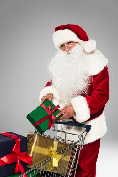 Santa claus in costume holding gift box near present in shopping cart isolated on grey — Stock Photo