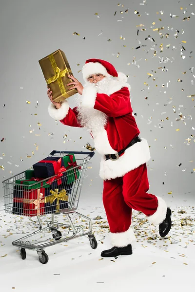 Santa Claus sosteniendo presente cerca del carrito de la compra y confeti sobre fondo gris - foto de stock