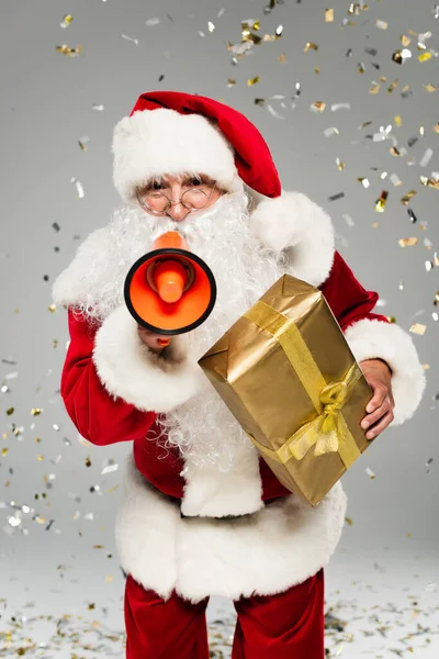 Père Noël en costume tenant cadeau et haut-parleur sous confettis sur fond gris — Photo de stock