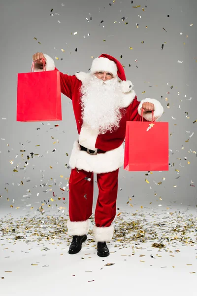 Santa claus holding red shopping bags under confetti on grey background — Stock Photo