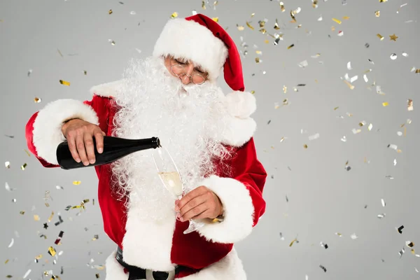 Санта-Клаус поливає шампанське під конфетті на сірому фоні — стокове фото